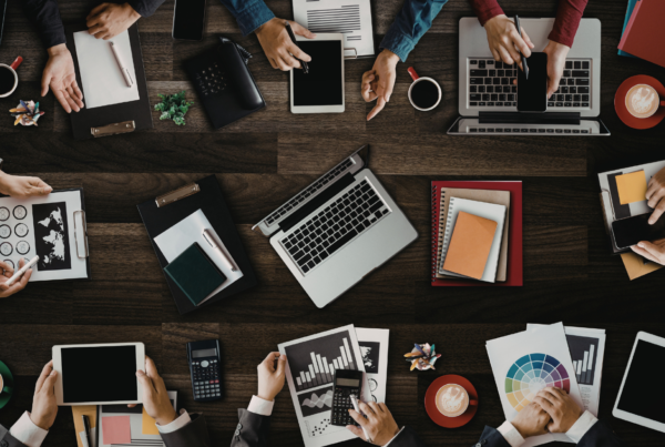 Widok z góry na duży, ciemny drewniany stół, przy którym wspólnie pracuje grupa ludzi. Widoczne są dłonie obsługujące laptopy, tablety, smartfony oraz analizujące dokumenty z wykresami. Zdjęcie ilustruje kreatywną przestrzeń do pracy i spotkań, charakterystyczną dla nowoczesnego biura lub środowiska typu coworking.