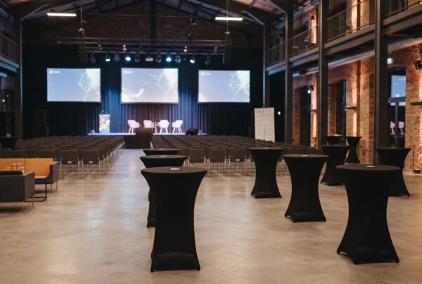 Wnętrze nowoczesnej sali konferencyjnej i strefy networkingowej w stylu industrialnym bez ludzi, z rzędami krzeseł, sceną i czarnymi stolikami koktajlowymi.